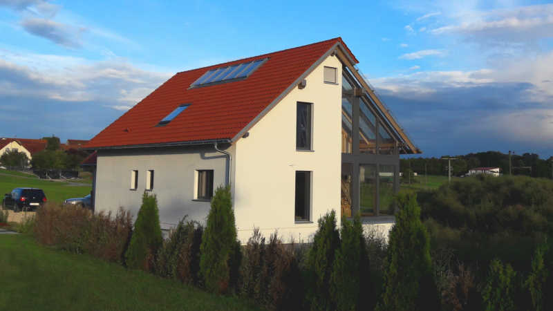 Kompaktes Satteldachhaus mit rotem Ziegeldach und verglastem Giebel in l&auml;ndlicher Umgebung.