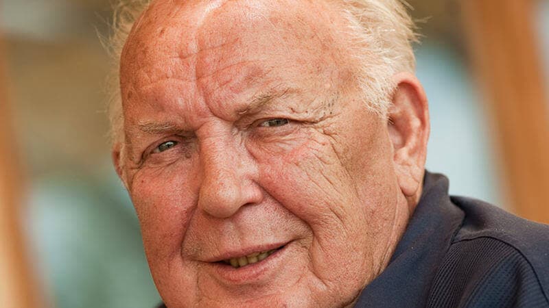 Klaus Becher, Portr&auml;t eines &auml;lteren Mannes mit grauen Haaren, freundlichem Blick und unscharfem Hintergrund.