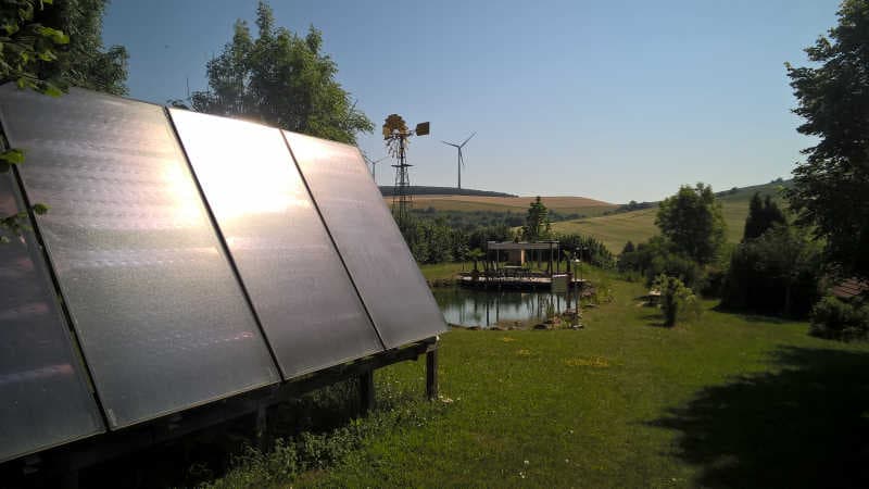 Solarkollektoren vor einem Naturteich; im Hintergrund Windr&auml;der und sanfte H&uuml;gellandschaft bei Sonnenschein.