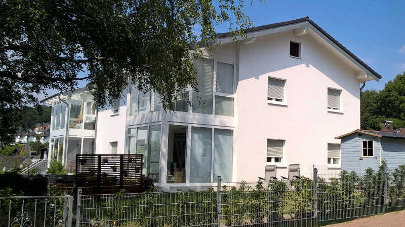 Modernes weißes Haus mit großflächiger Verglasung und Balkon, Zaun und Bäume im Vordergrund.