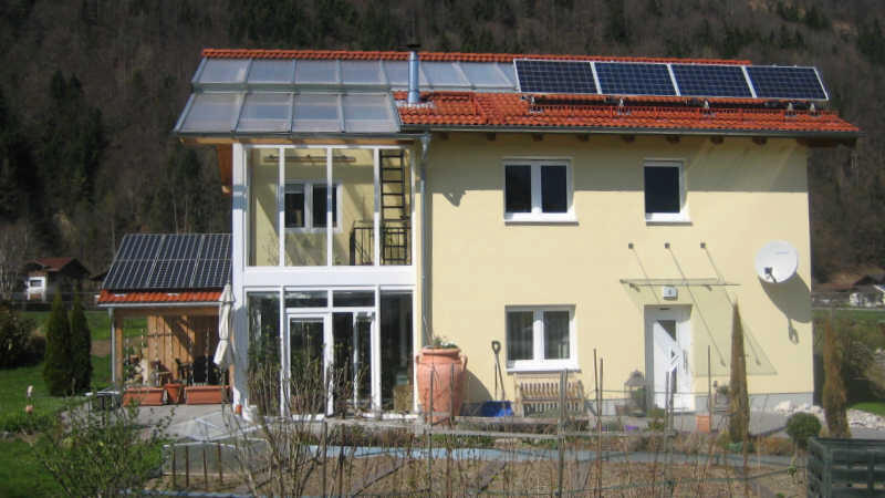 Gelbes Einfamilienhaus mit Wintergarten/Glasanbau und Photovoltaik auf dem Dach, Rückansicht.