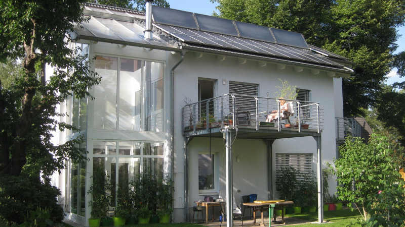 Modernes weißes Haus mit großer Glasfront und Balkon, Gartenmöbel auf der Terrasse.
