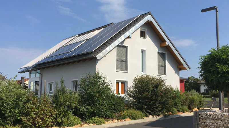 Weißes Einfamilienhaus mit Satteldach und Vorgarten, sonnige Außenansicht.