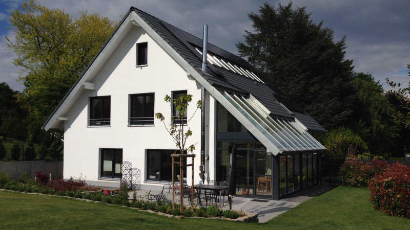 Weißes Einfamilienhaus mit Satteldach, PV-Anlage und verglastem Wintergarten, Gartenanlage davor.