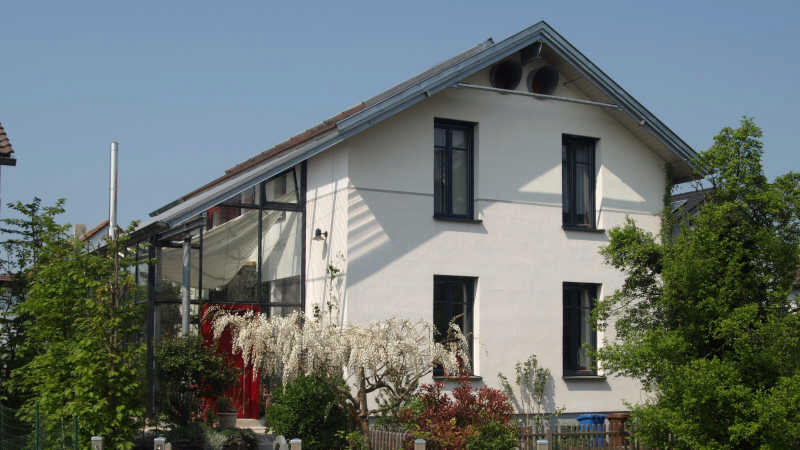 Weißes Einfamilienhaus mit Glaswintergarten an der Seite, Garten mit Blumen im Vordergrund.
