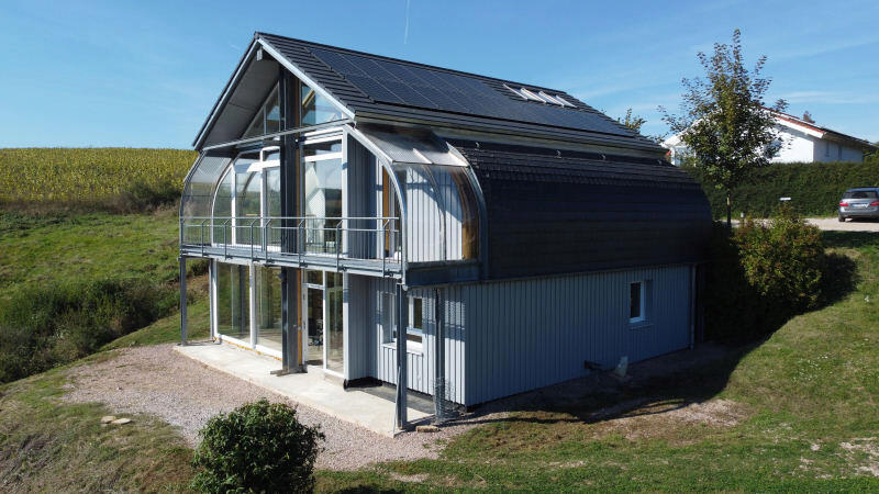 Langes Solarhaus mit schrägem Dach, kombiniert aus PV-Modulen und Solarthermie, umgeben von Bäumen.