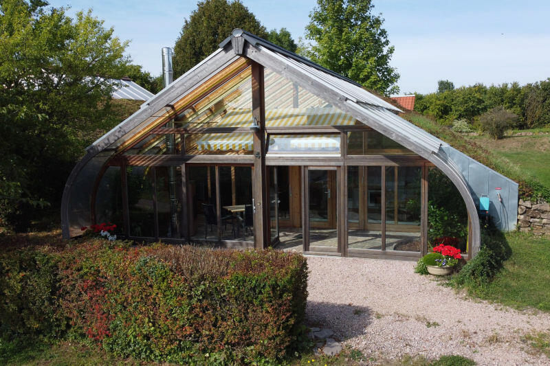Solarhaus mit gewölbter Glasfassade und Metallbögen, Wintergarten vorn, Hecken und Kiesweg im grünen Garten.