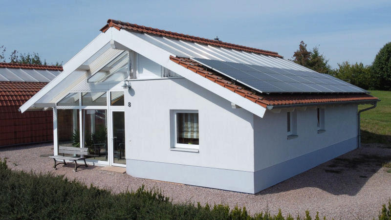 Weißes Solarhaus im Bungalowstil mit Satteldach und großer Photovoltaikanlage, Terrasse mit Sitzbank.