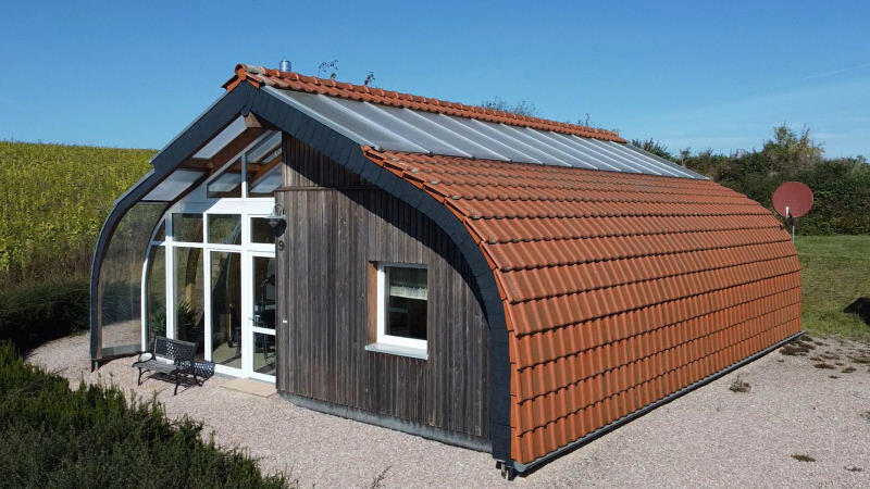 Kompaktes Holz-Solarhaus mit geschwungener Ziegeldachhaut und verglaster Eingangsfassade, ländliche Lage.